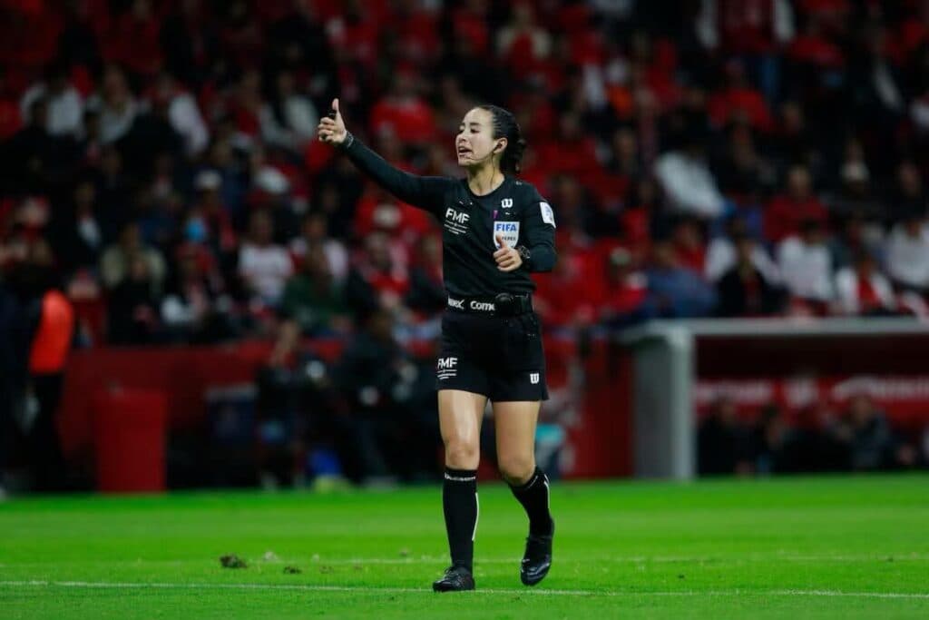Katia Itzel García, árbitra mexicana, dirigiendo un partido de fútbol con gesto de aprobación en un estadio lleno de espectadores.