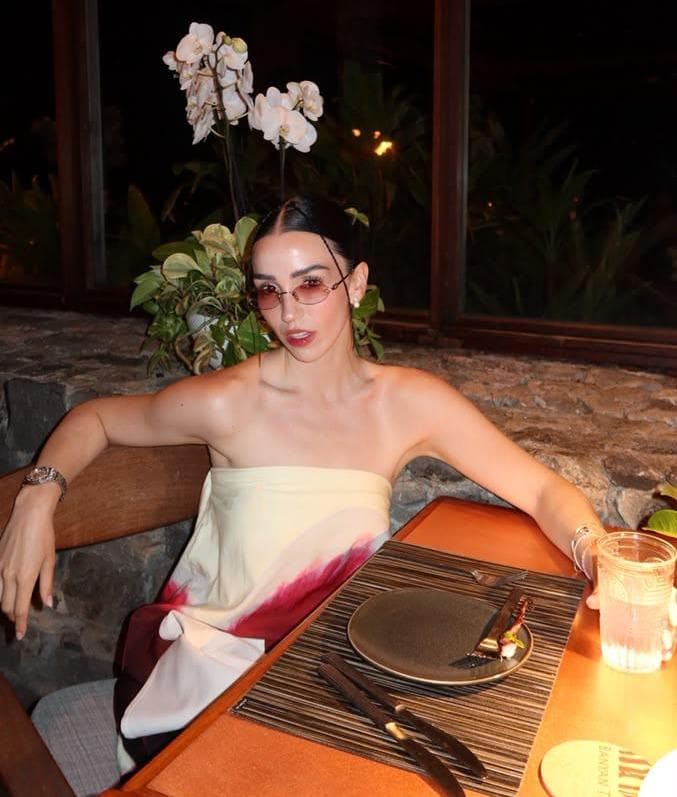 Estíbaliz Badiola posando en un restaurante, luciendo un vestido sin mangas y gafas de sol, rodeada de plantas y decoración elegante.