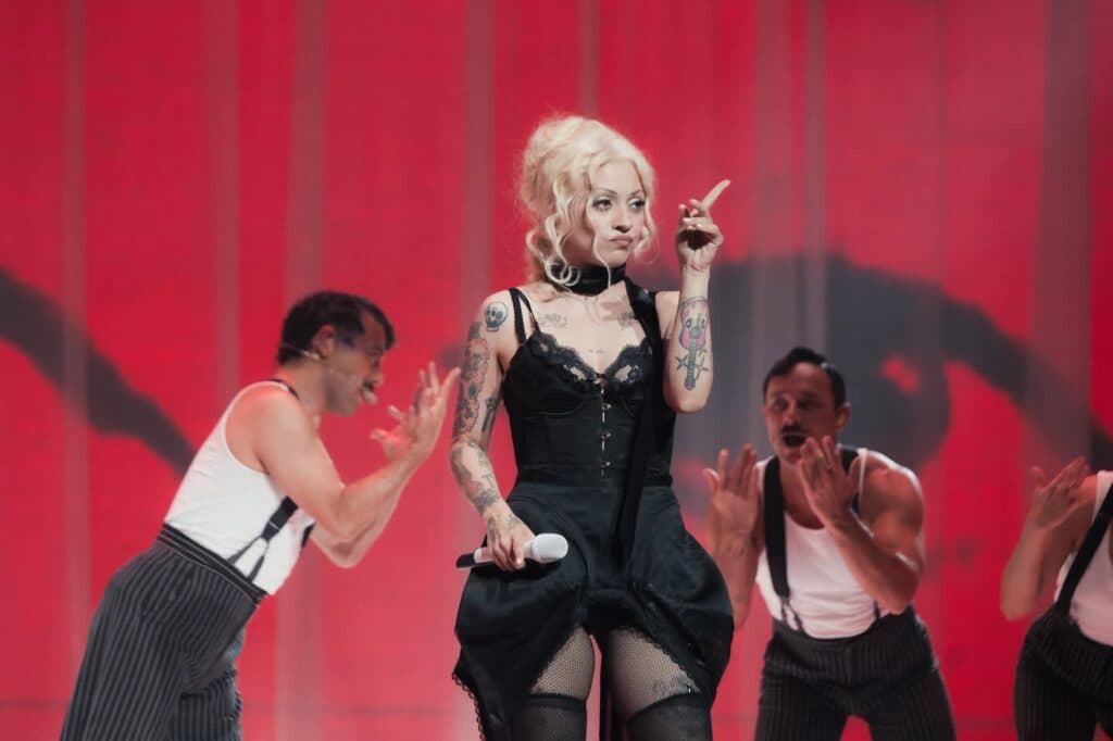 Mon Laferte en el escenario durante una actuación, luciendo un vestido negro y rodeada de bailarines con vestimenta de rayas.