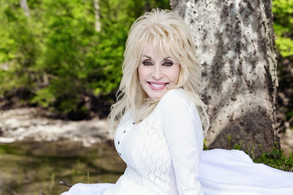Dolly Parton sonríe sentada al aire libre, rodeada de naturaleza, en un tributo a su legado en la música country.