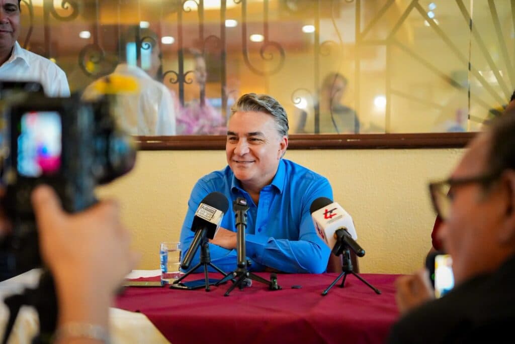 Jesús Ruiz Uribe sonríe durante una conferencia de prensa sobre las asambleas de MORENA en Baja California, con micrófonos y cámaras alrededor.