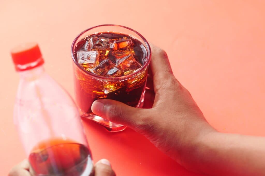 Un vaso con refresco y hielo, mientras una mano sostiene una botella de bebida azucarada, resaltando el riesgo para la salud del hígado.