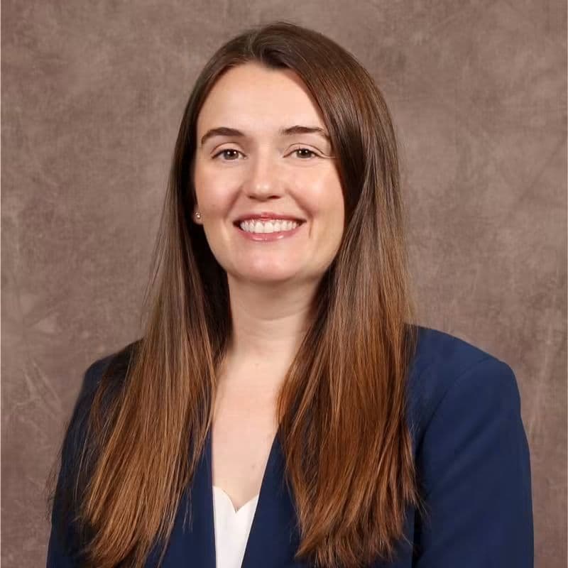 Mujer sonriente con cabello largo y lacio, vestida con un blazer oscuro, en un fondo neutro, relacionada con el caso de