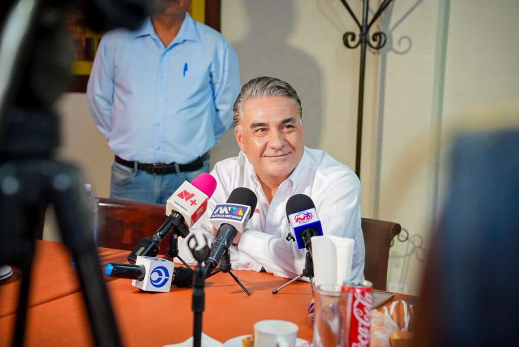 Jesús Ruiz Uribe sonríe durante una conferencia de prensa, rodeado de micrófonos y con un ambiente de diálogo sobre MORENA en Tijuana.
